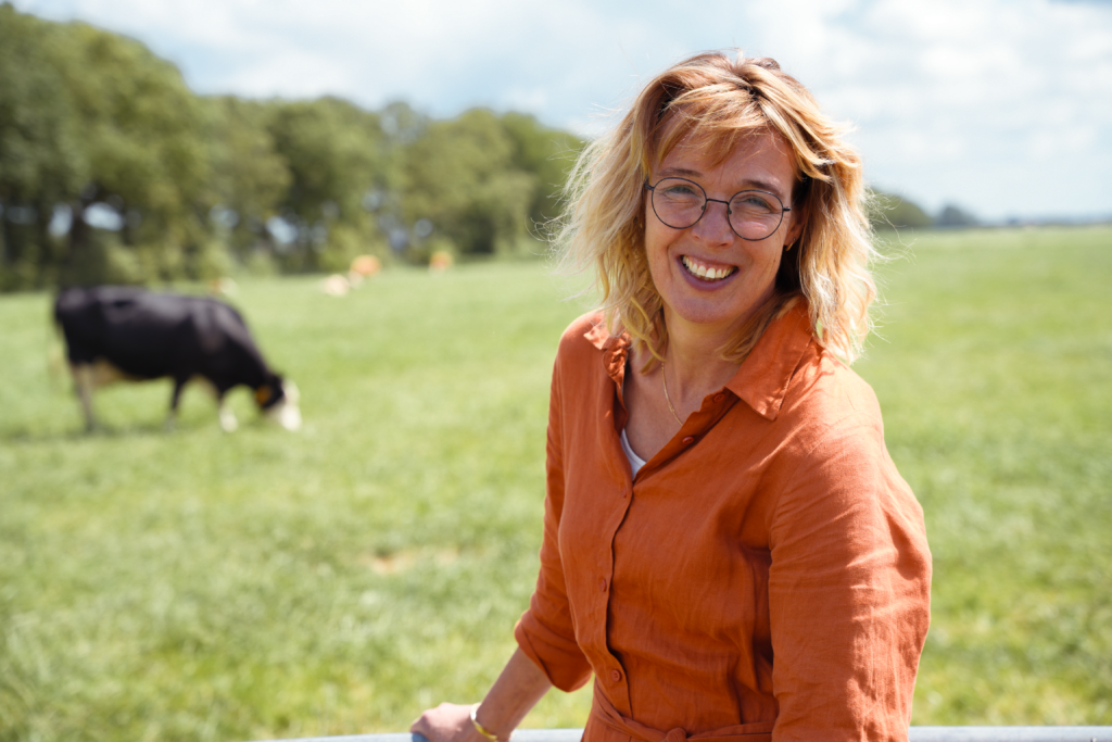 Nynke Benedictus. Een vrouw met blond haar en een bril en een oranje blouse. Op de achtergrond een weiland met een grazende koe.