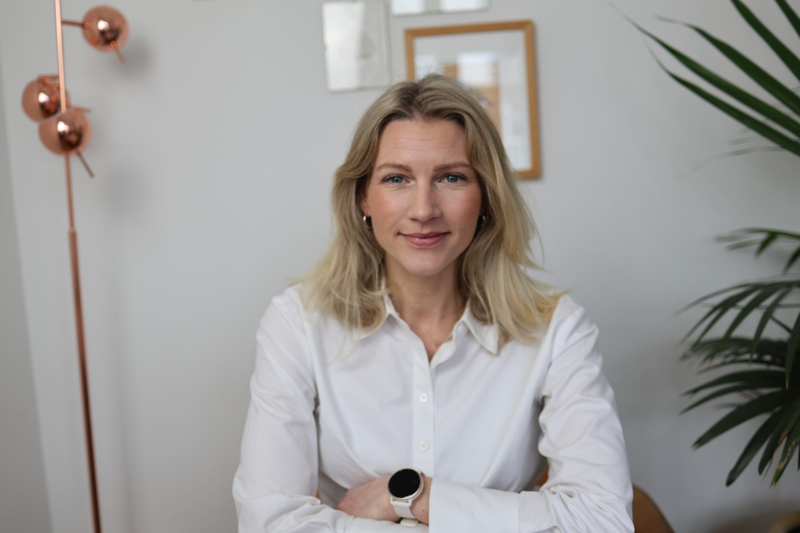 Een vrouw met blond haar en een witte blouse. Haar armen over elkaar met een witte smartwatch te zien. Op de achtergrond een staande lamp in koperen kleur.
